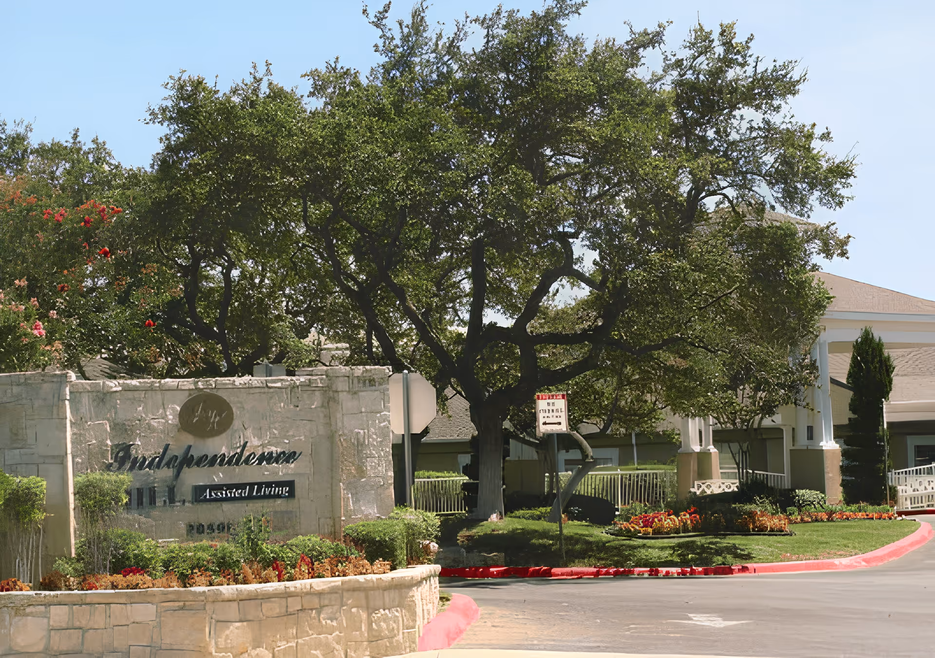 Entrance sign and landscaped front of the Independence Village assisted living facility with a large tree and driveway.