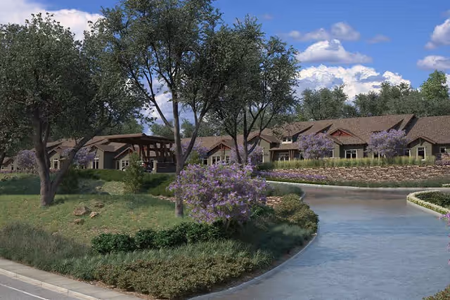 Front exterior of a single-story senior living building set behind landscaped lawns, trees, purple-flowering shrubs, and a curved waterway or driveway under a blue sky.