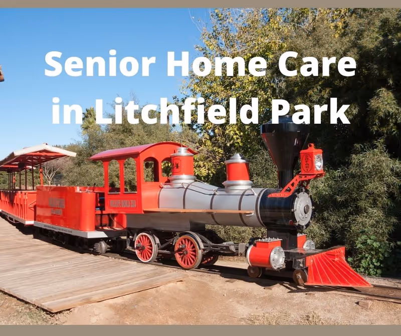 A bright red miniature train runs on outdoor tracks beside trees and a wooden platform.