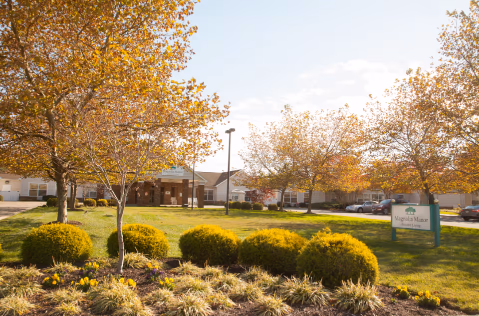 Front exterior of a senior living facility with landscaped lawn, trees, and a 'Magnolia Manor' sign.