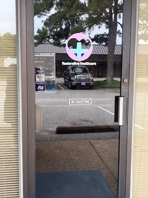 Glass door with pink heart logo reading Restorative Healthcare, reflecting parking lot and building exterior.