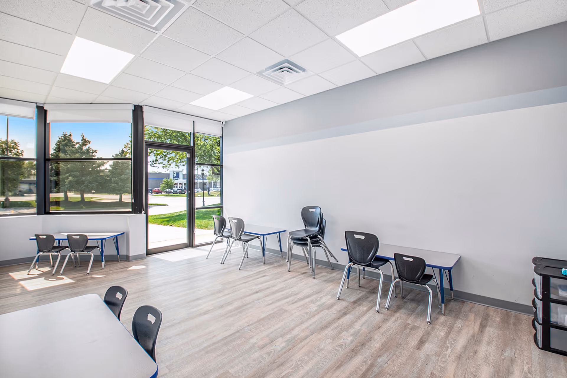 Bright, empty classroom-like interior with large windows, a door, and stacked chairs along walls.