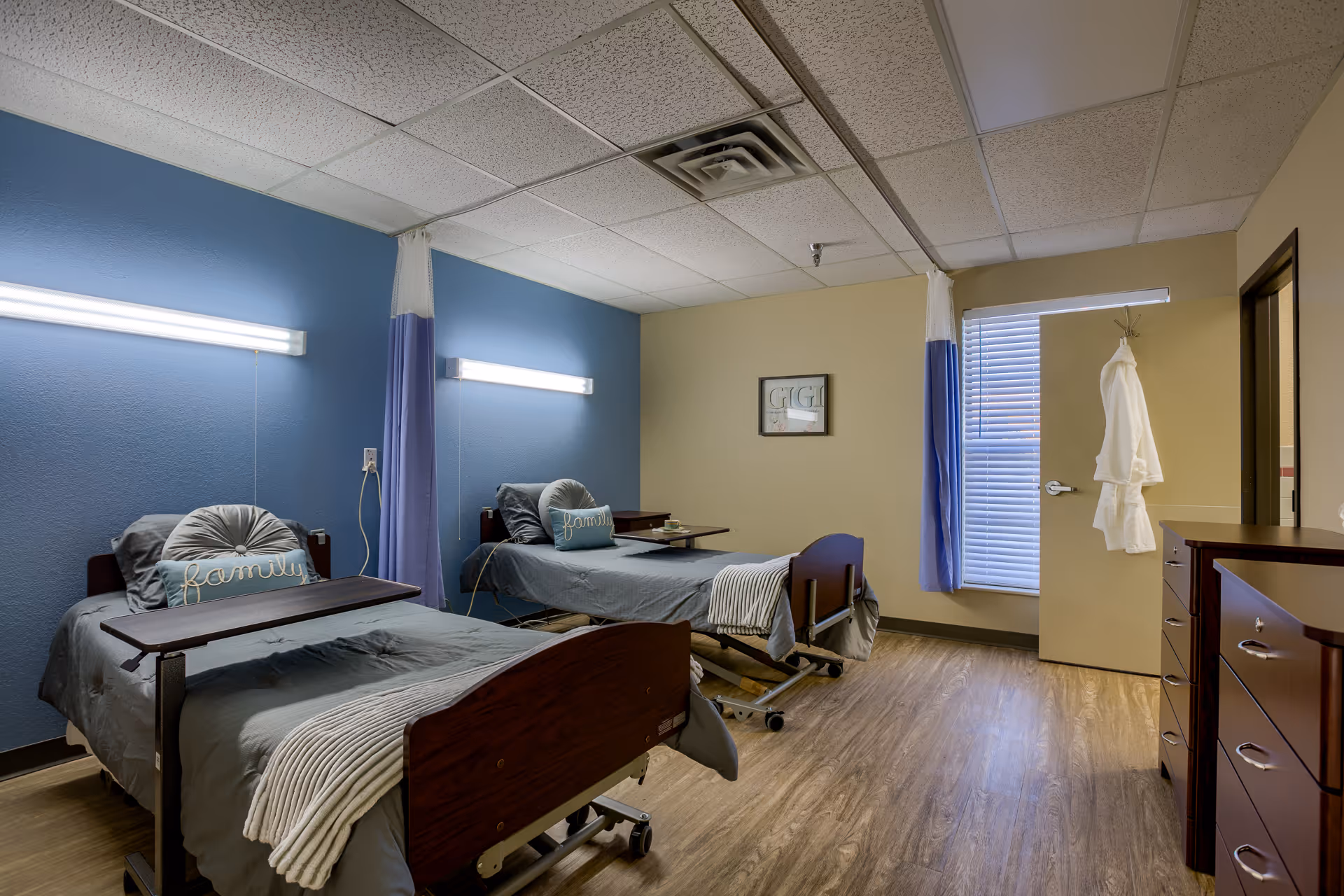 A room with two hospital-style beds separated by a curtain. Each bed has gray bedding, a round pillow, and a pillow with the word 'family' embroidered on it. There are wall-mounted lights above each bed, a window with blinds, a door with a white robe hanging on the handle, and a wooden dresser on the right side. The walls are painted blue and beige, and the floor is wood-patterned.