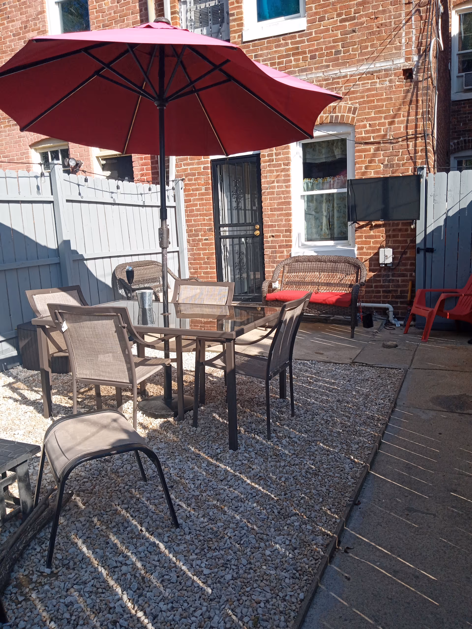 Brick building backyard patio with gravel ground, glass table, chairs, and a red umbrella.