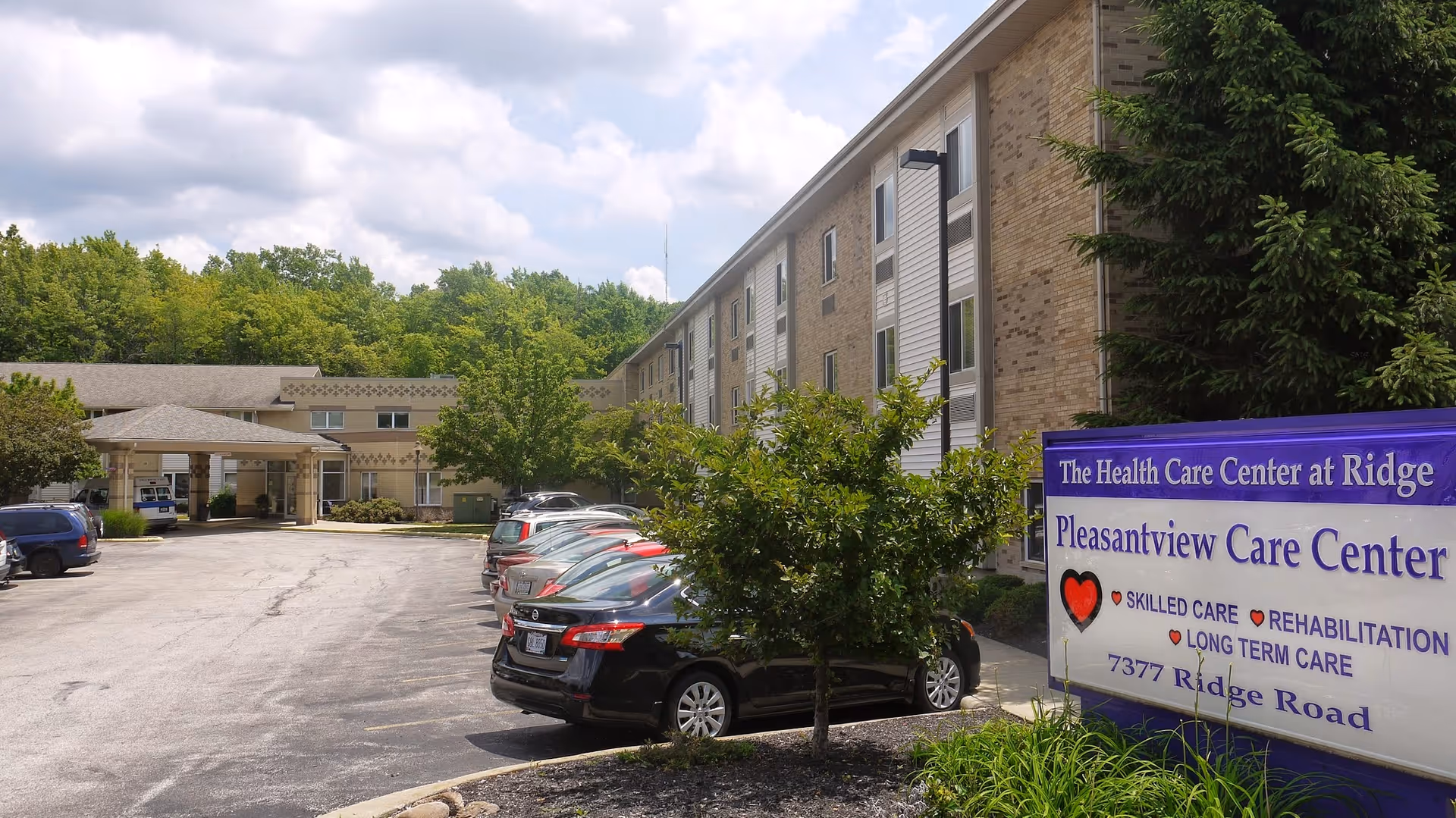 Exterior view of Pleasantview Care Center showing a multi-story brick building with several parked cars in front. There is a sign in the foreground that reads 'The Health Care Center at Ridge Pleasantview Care Center' along with services offered and the address 7377 Ridge Road. Trees and greenery surround the parking area under a partly cloudy sky.