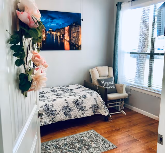 Cozy bedroom in a senior facility featuring a bed, chair, window, and floral decor.