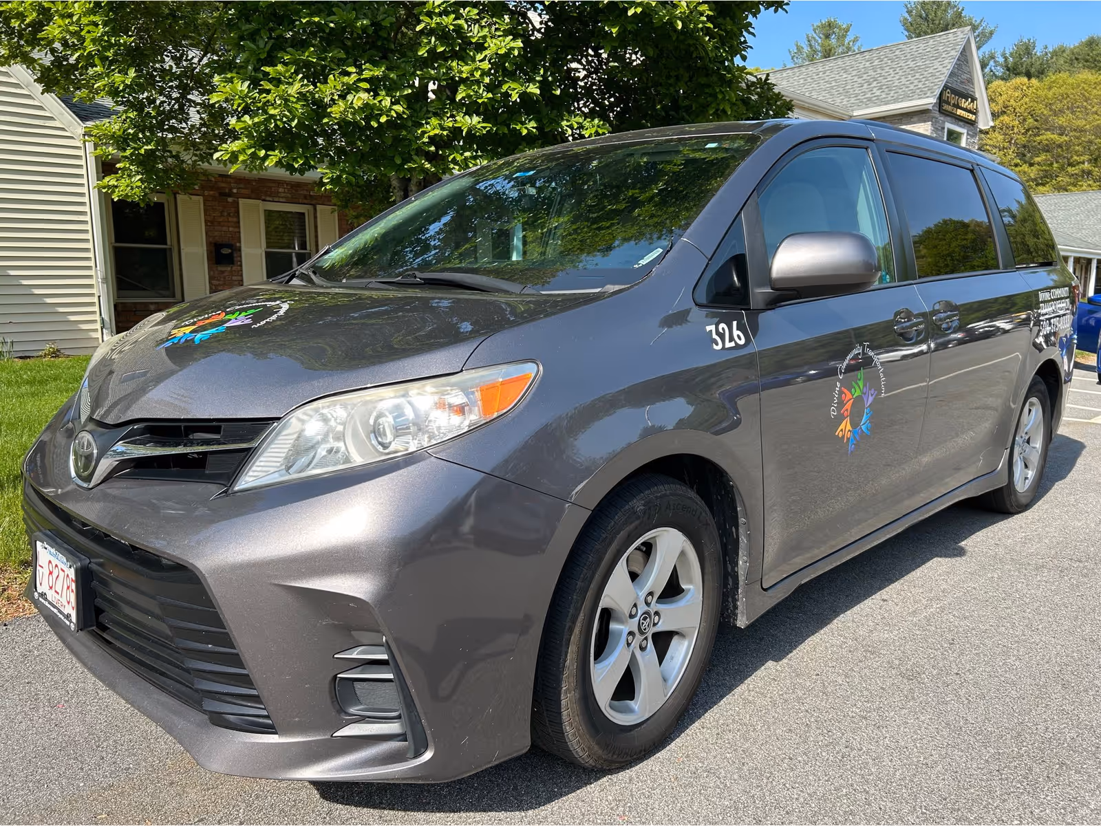 Grey passenger van with a colorful logo parked outside a residential building under trees.