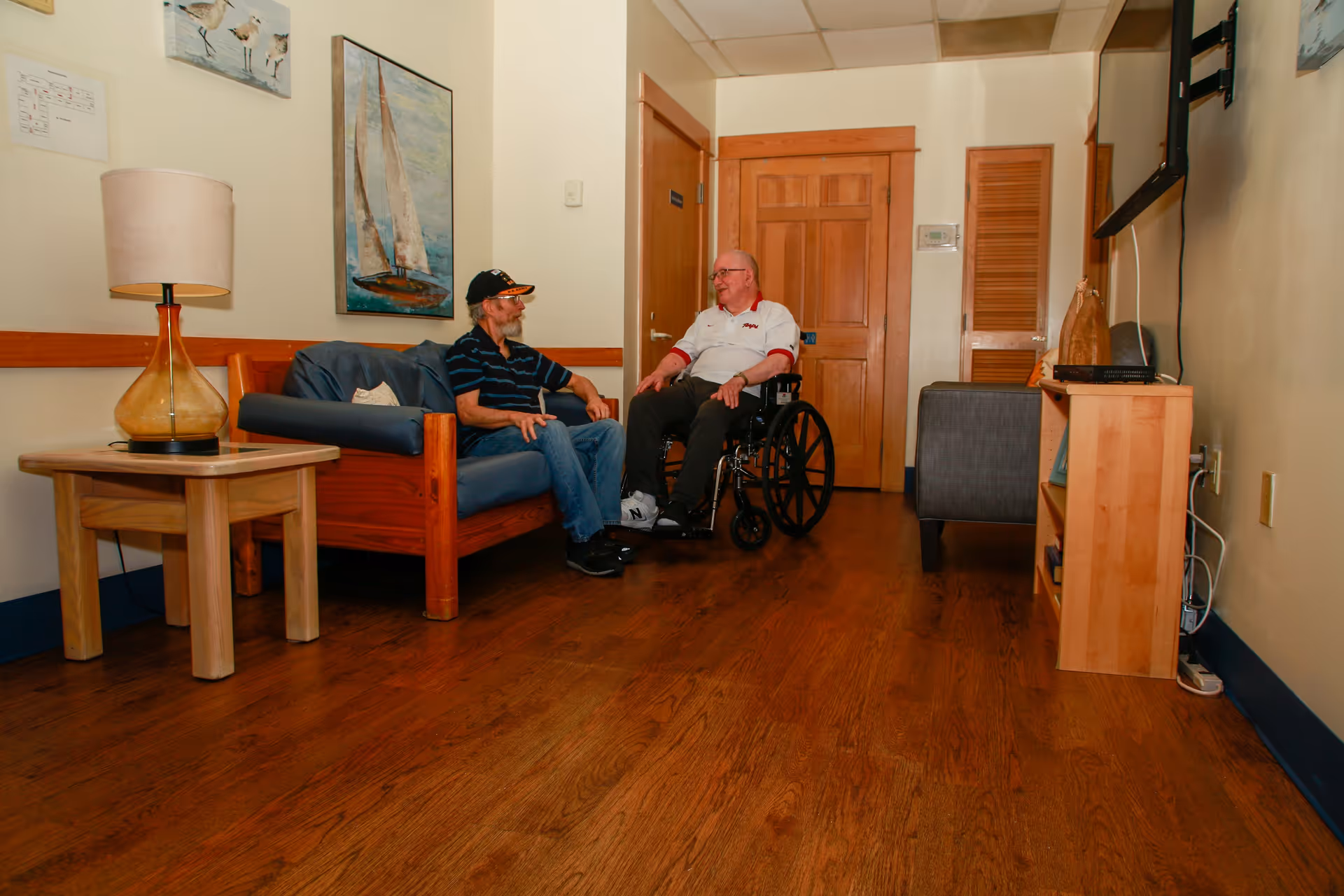 Two elderly men, one in a wheelchair, talking in a wood-floored sitting area with a sofa, lamp, artwork, and a wall-mounted TV.