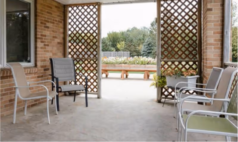 Covered patio with several chairs and lattice screens opening onto a garden with planters and trees.