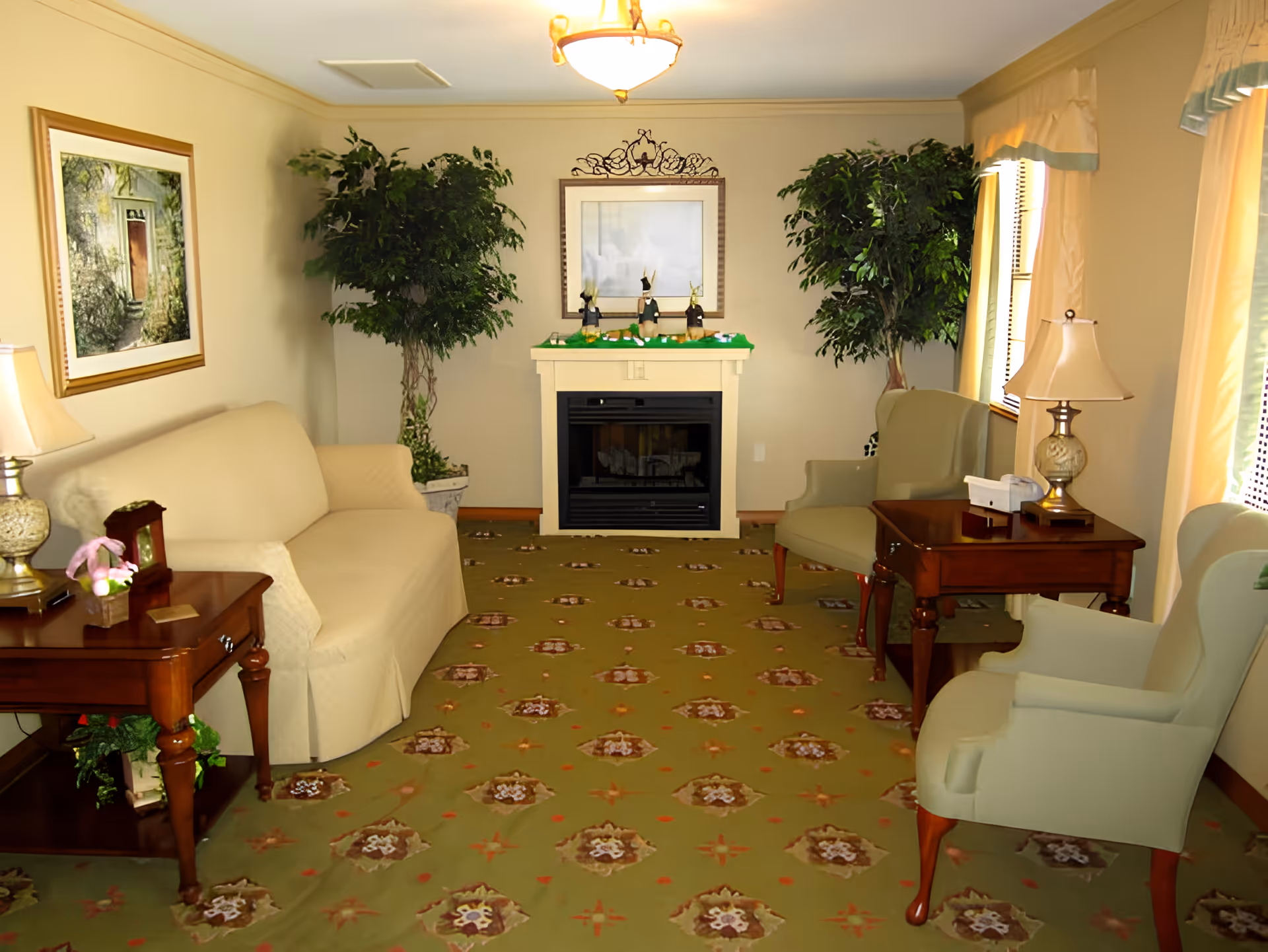 A cozy living room with a beige sofa on the left and two light green armchairs on the right. There are two wooden side tables with lamps on either side of the room. The room features a patterned green carpet, two large potted plants flanking a white fireplace with decorative items on the mantel, and a framed picture above the fireplace. Light filters in through windows with cream-colored curtains on the right side.