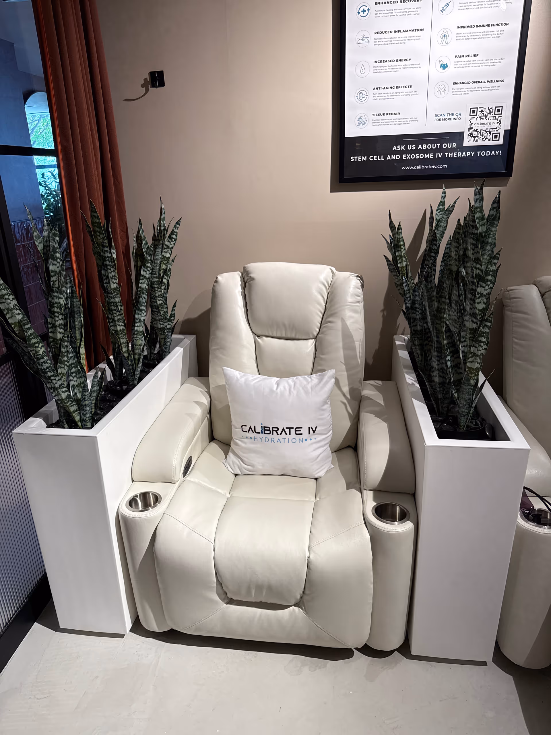 A white reclining chair in a modern lounge with tall planters and a Calibrate IV poster.