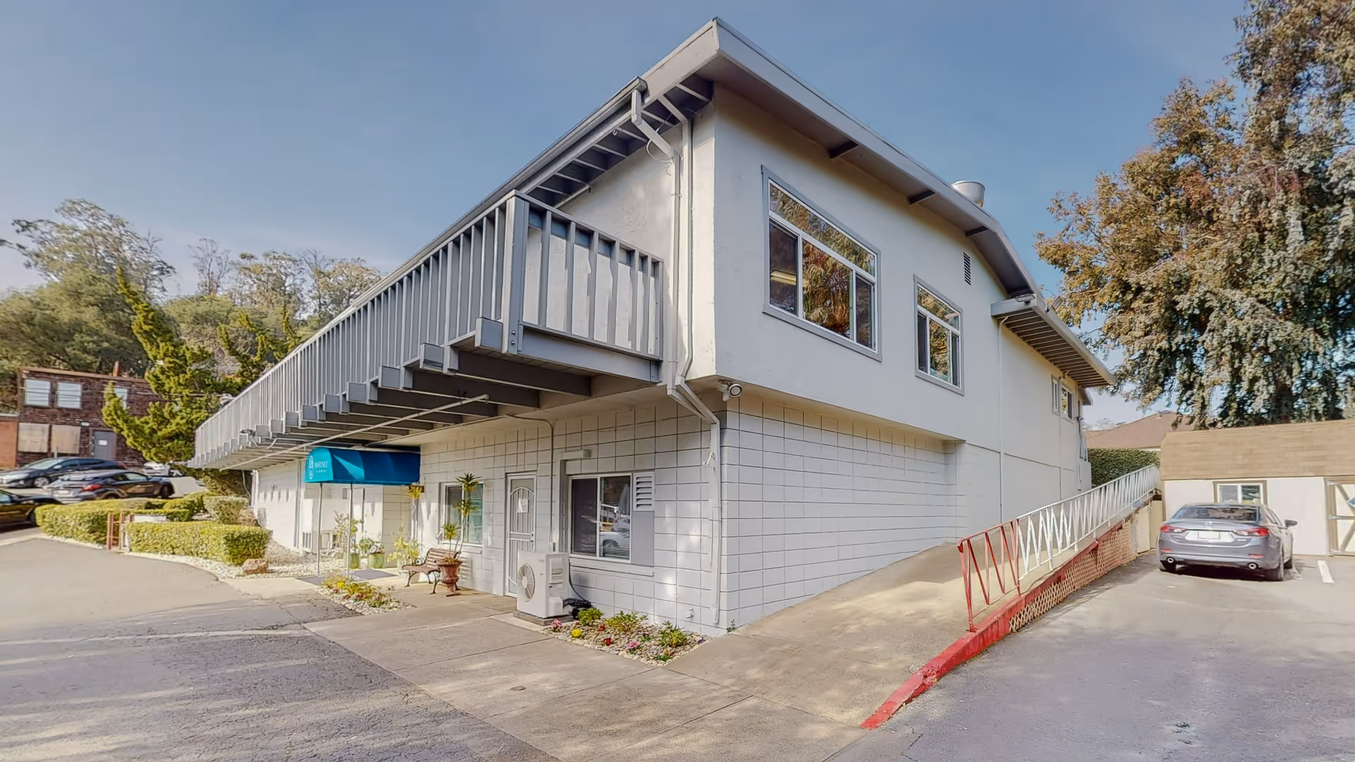 Two-story light-gray healthcare center exterior with a ramp, parking, and surrounding trees.