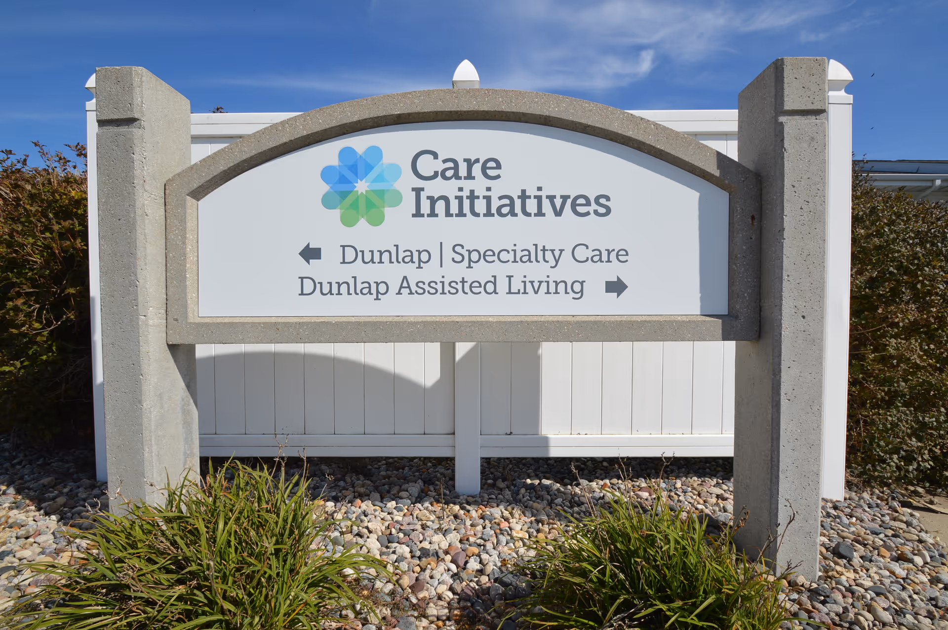 Outdoor sign for Care Initiatives with directions to Dunlap Specialty Care and Dunlap Assisted Living, set against a white fence and surrounded by rocks and greenery under a clear blue sky.