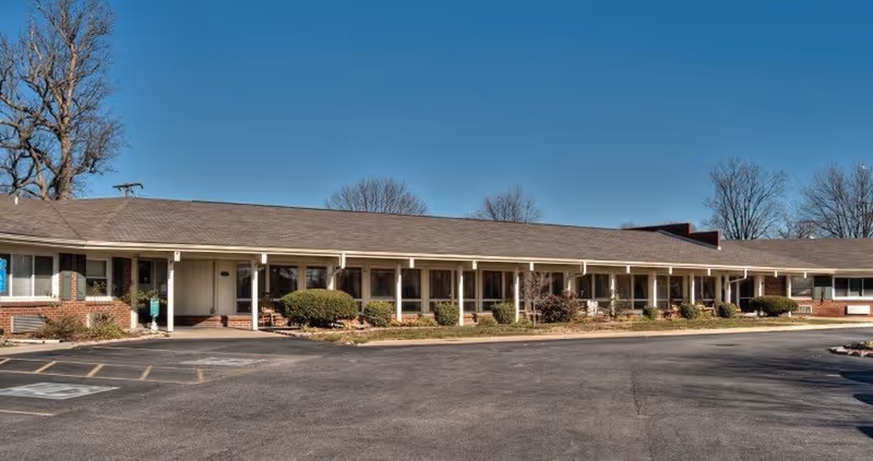 One-story brick and siding senior living building with a covered front porch, shrubs, and an empty parking lot under a clear blue sky.