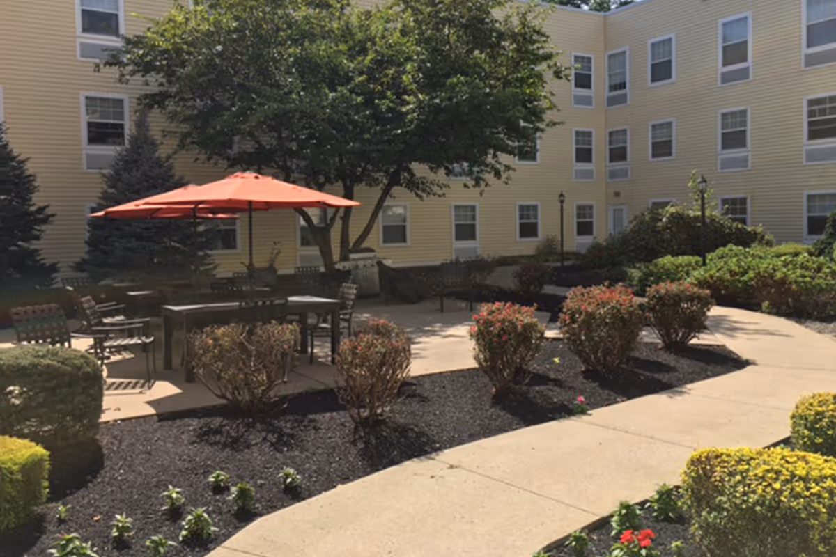 Outdoor courtyard with a red umbrella, tables and chairs amid landscaped beds in front of a yellow multi-story residential building.