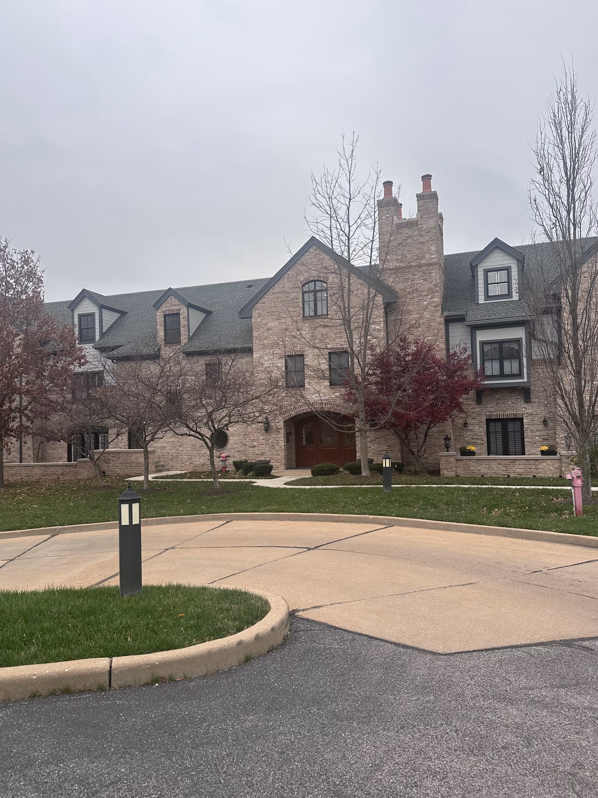 Brick multi-story senior living building with dormer windows, arched main entrance, trees, and a circular driveway in front.