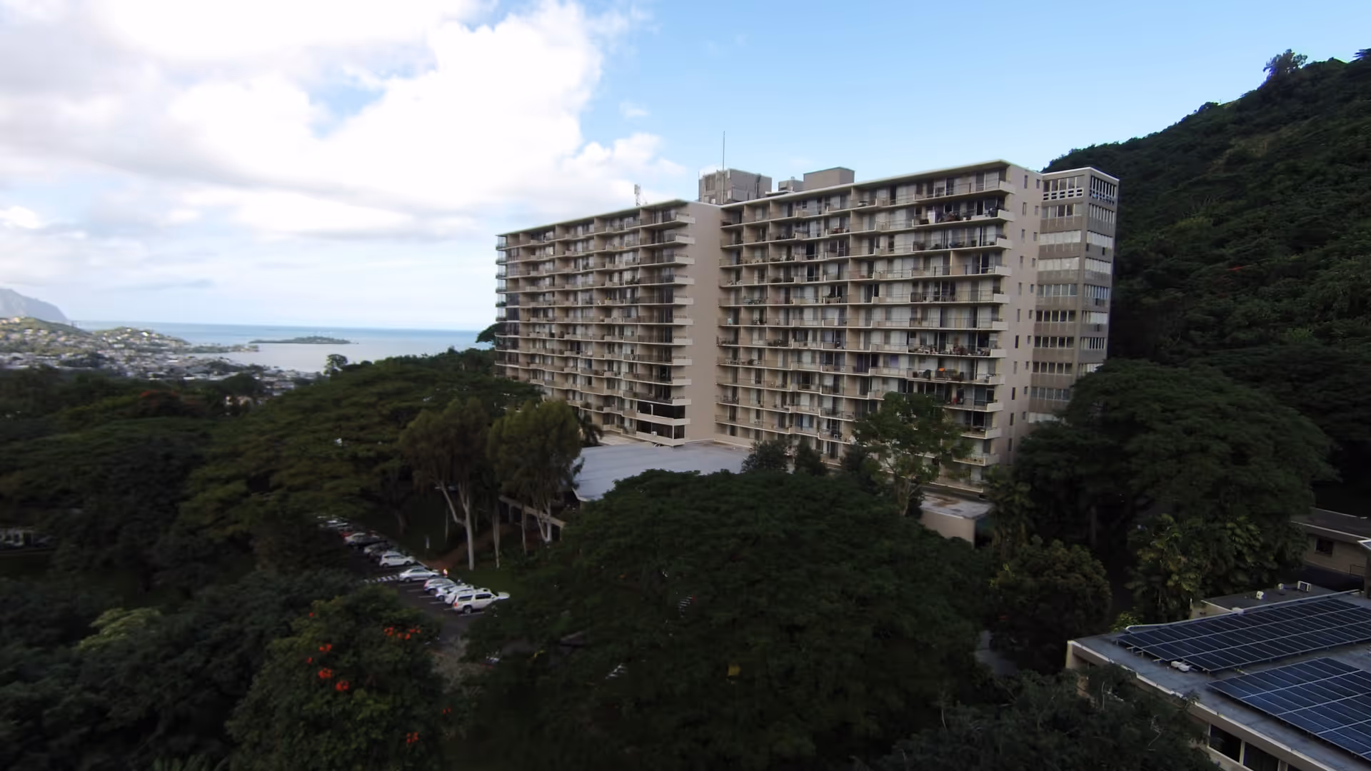 A large multi-story residential building surrounded by trees with hills and ocean in the background.