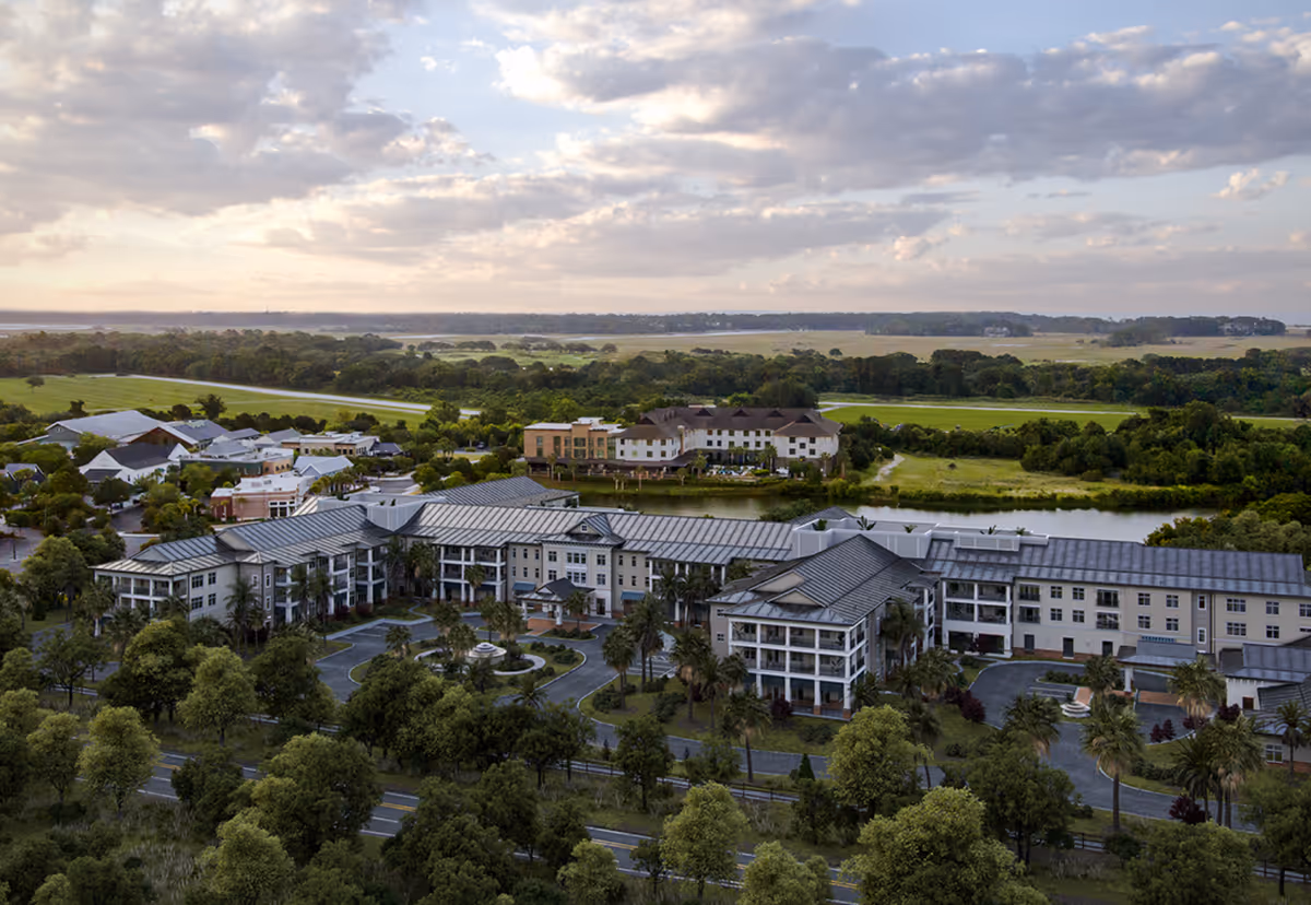 Aerial view of a large senior living complex with landscaped grounds, a central fountain, and surrounding marshland under a cloudy sky.