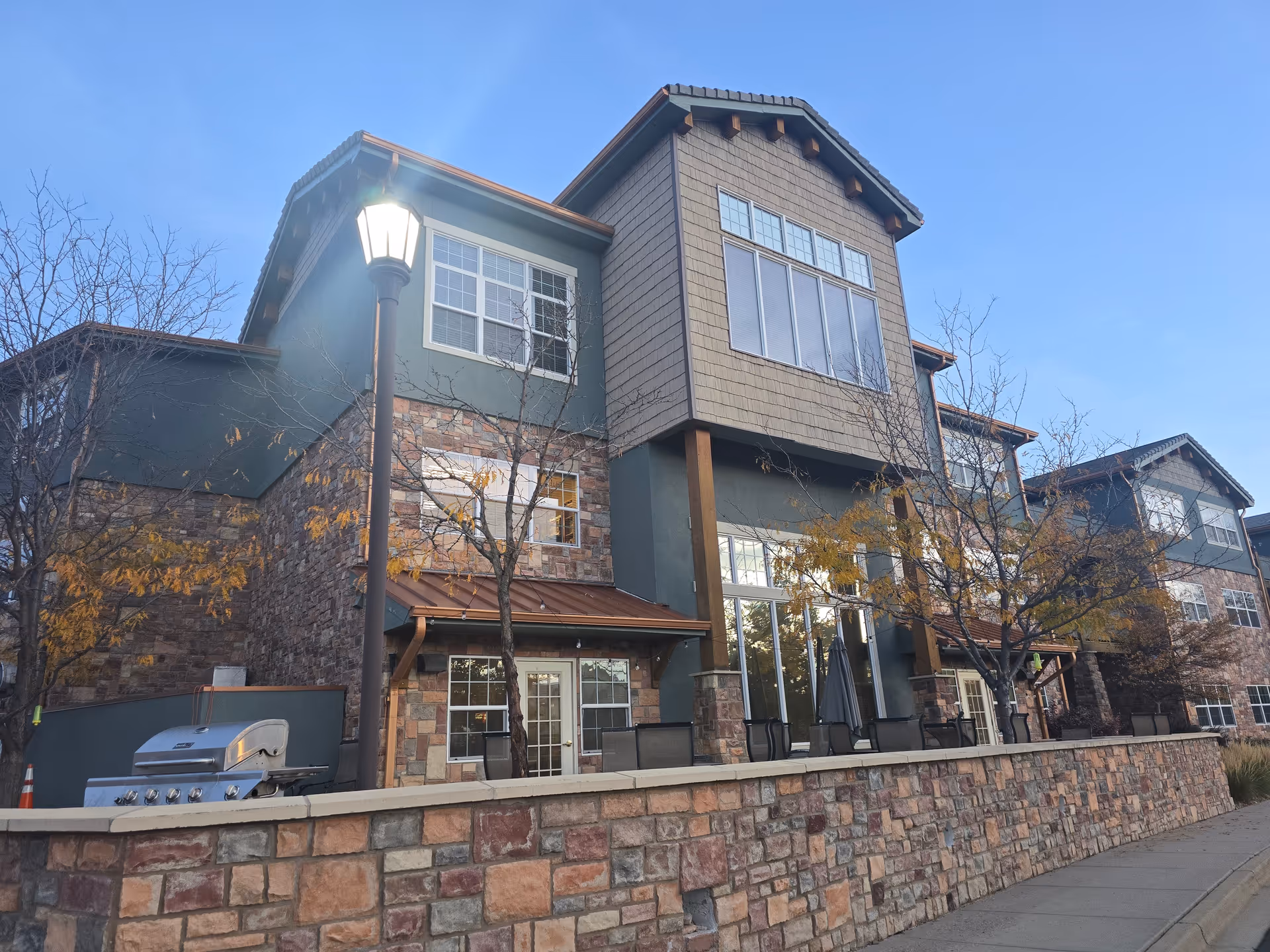 Exterior view of a multi-story senior living facility building with stone and green siding, large windows, a patio area with chairs and umbrellas, and a street lamp in front. Trees with autumn leaves are visible around the building under a clear blue sky.