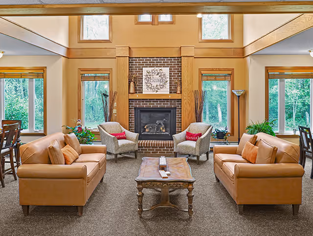 Cozy senior living lounge with brick fireplace, tan sofas, chairs, and a wooden coffee table.