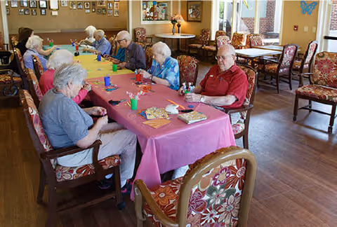 Elderly residents seated around a long table with a pink tablecloth in a communal dining/activity room.