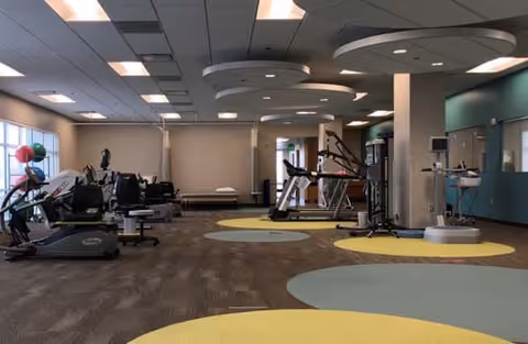 A spacious fitness room with exercise equipment including stationary bikes, treadmills, and weight machines. The room has a carpeted floor with circular colored patterns in yellow and blue, large windows on the left side, and ceiling lights with circular fixtures.
