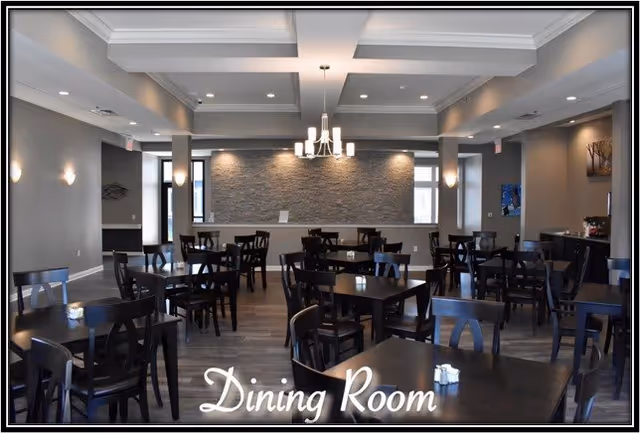 A spacious dining room with multiple dark wooden tables and chairs arranged neatly. The room features a modern chandelier hanging from a coffered ceiling, recessed lighting, and a textured stone accent wall at the far end. Large windows allow natural light to enter, and the walls are painted in neutral tones.