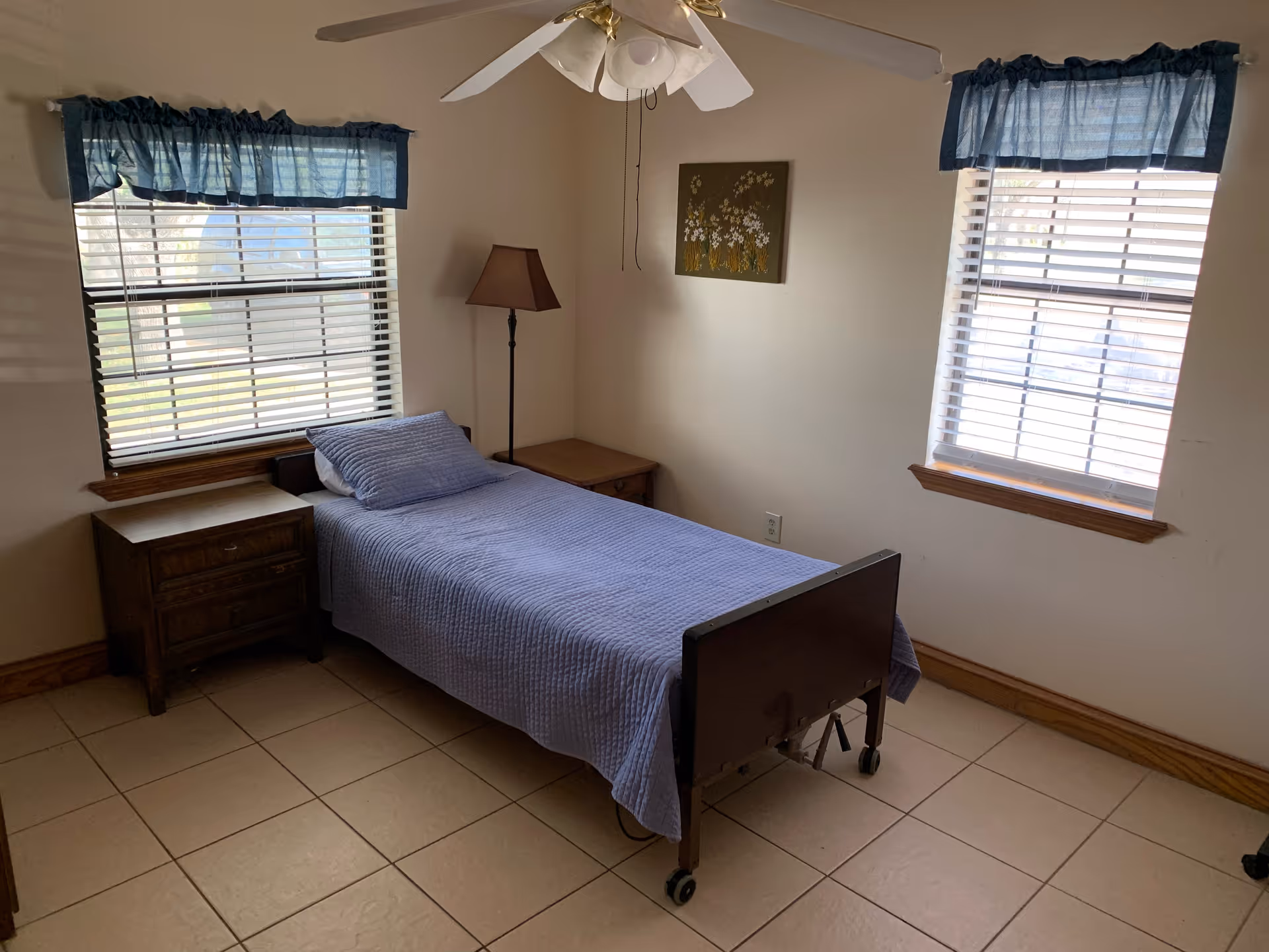 A small bedroom with a single bed covered in a blue quilt and matching pillow. The bed has wheels and is positioned between two windows with blue valances. There is a wooden nightstand on each side of the bed, and a floor lamp with a brown shade is placed next to the bed. A ceiling fan with lights is mounted on the ceiling, and a small floral painting hangs on the wall above the bed.