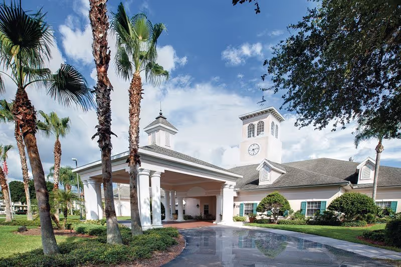Sunny exterior view of Aston Gardens Sun City Center with porte-cochère entrance and clock tower.