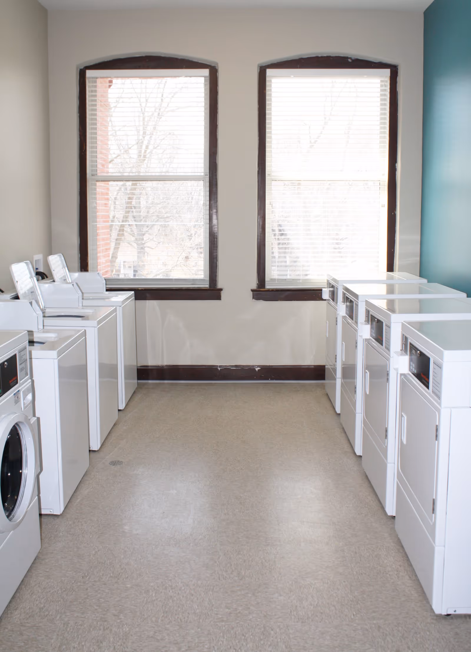 A bright laundry room with white washing machines and dryers lined along both walls and two windows at the far end.