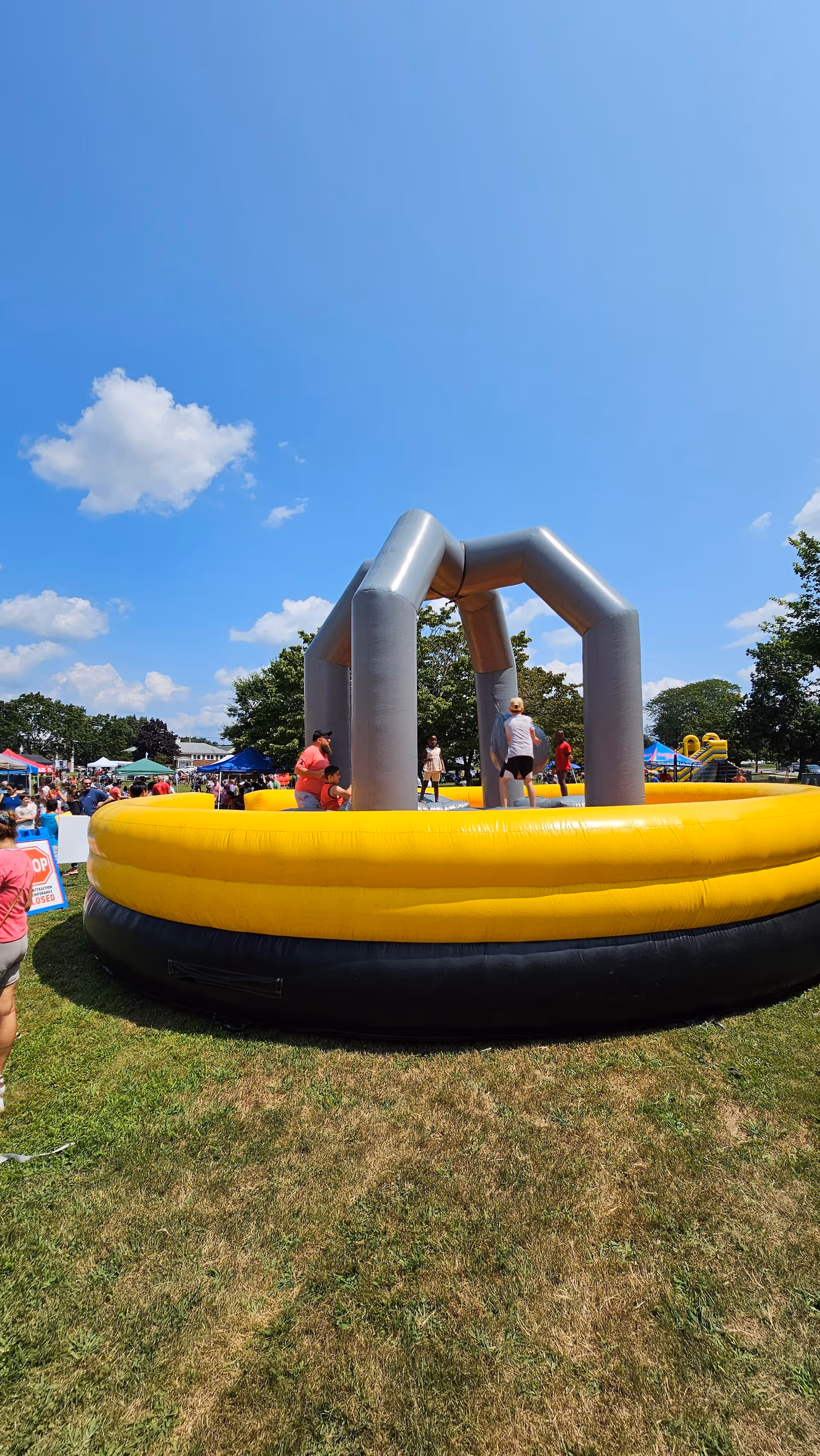 Outdoor inflatable obstacle course in yellow and gray, with people around on a grassy field.