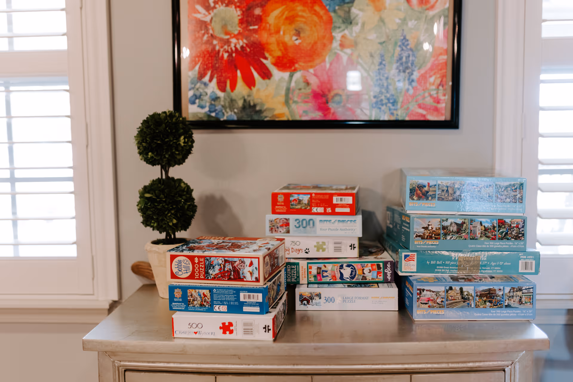 Table with stacked puzzle boxes and a small topiary in front of colorful wall art.