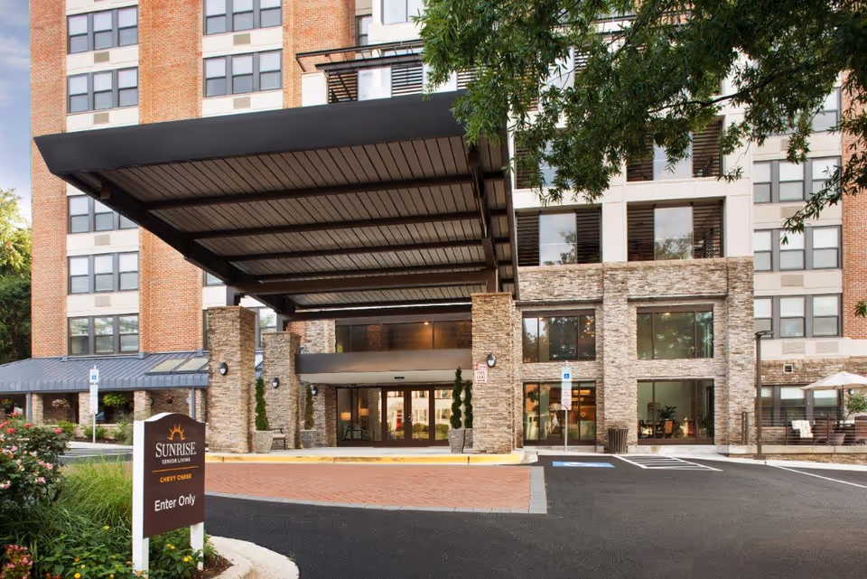 Front entrance of the Sunrise of Chevy Chase senior living building showing a covered drop-off porte-cochère, glass doors, and a landscaped drive with a 'Sunrise' sign.