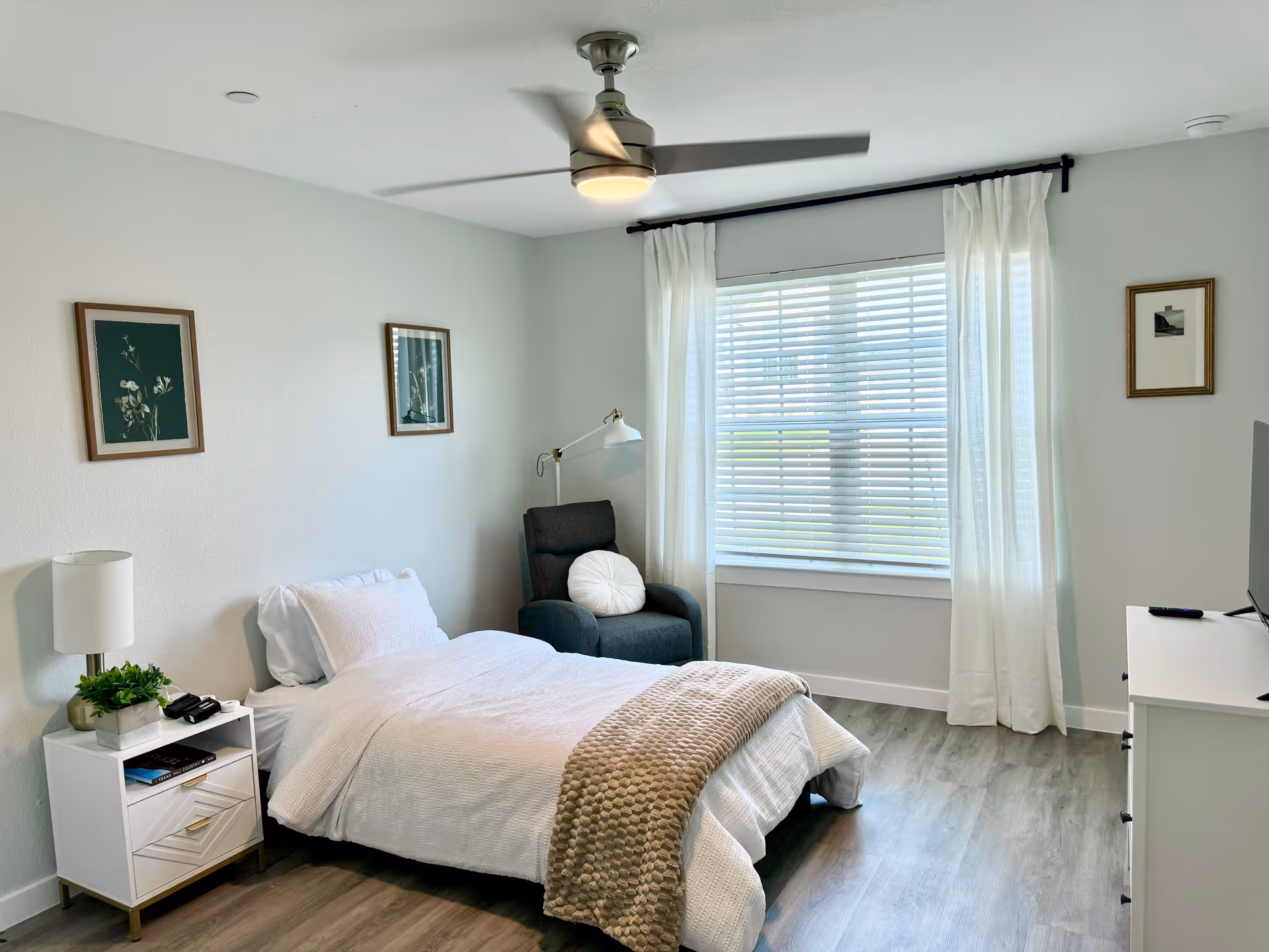A bright, modern bedroom with a bed, chair, window, and dresser in soft tones.