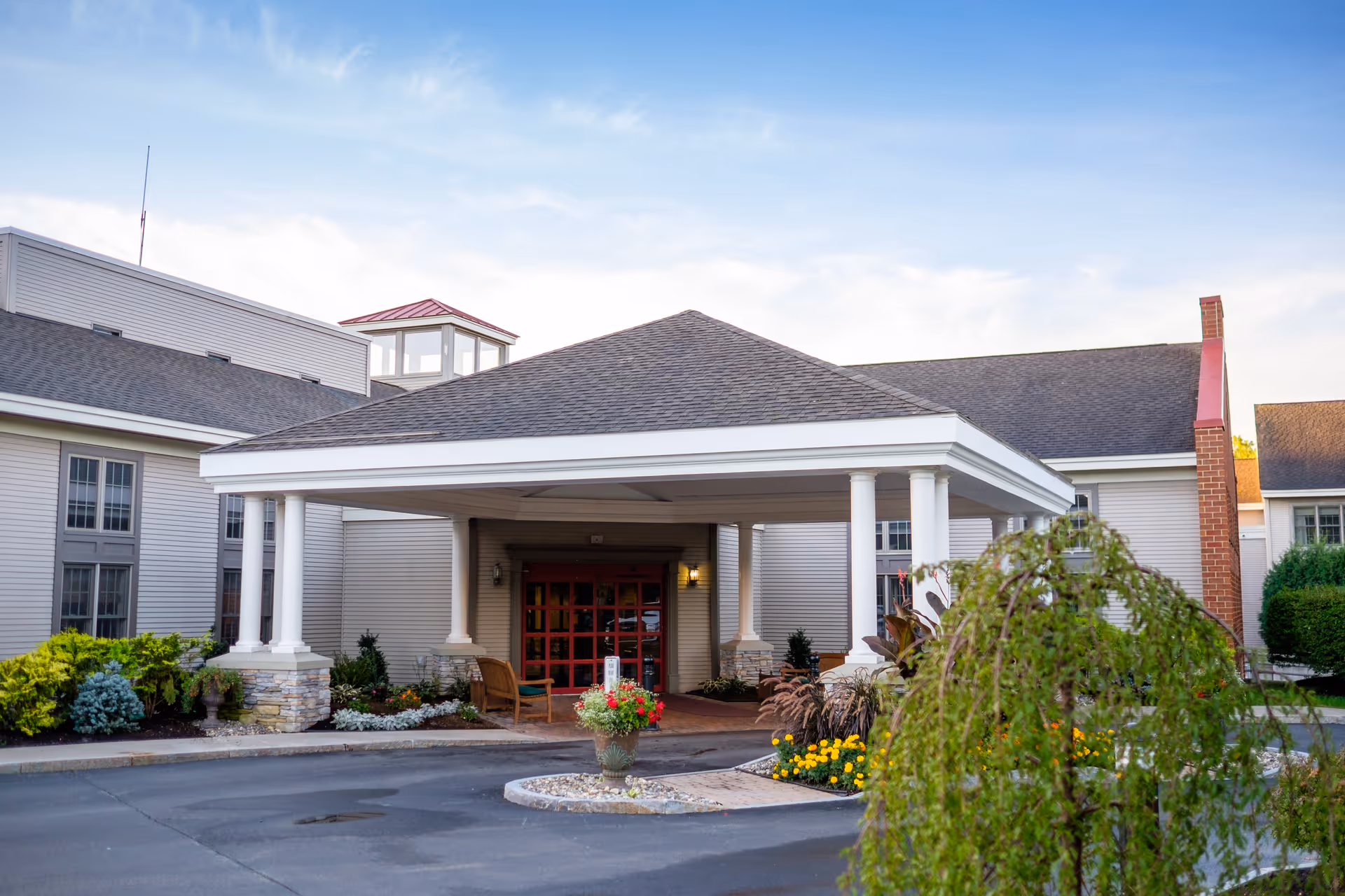 Entrance of a senior living facility with a covered driveway supported by white columns, surrounded by landscaped greenery and flowers under a partly cloudy sky.