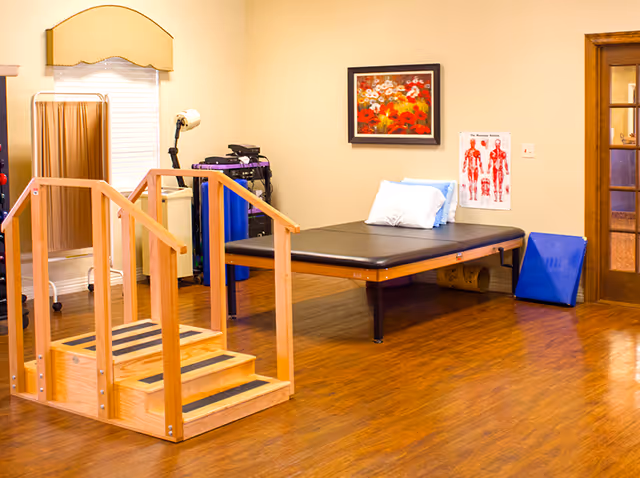 A rehabilitation therapy room with a wooden step exercise apparatus in the foreground, a black padded therapy table with pillows against the wall, a medical chart showing muscle anatomy, a framed floral painting, and various therapy equipment in the background.
