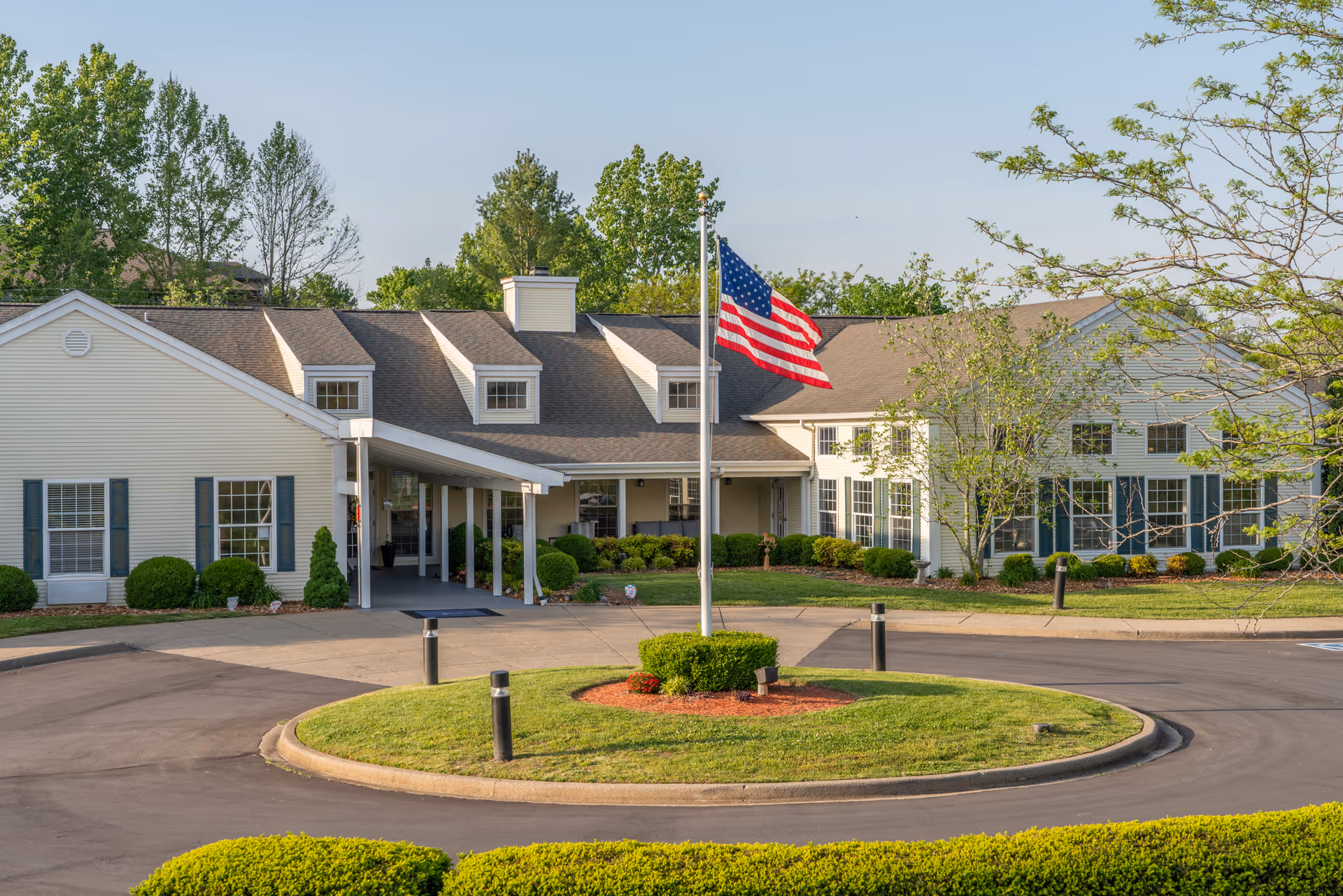 Front entrance of a single-story senior living building with a covered drop-off, circular driveway, landscaping, and an American flag on a flagpole.