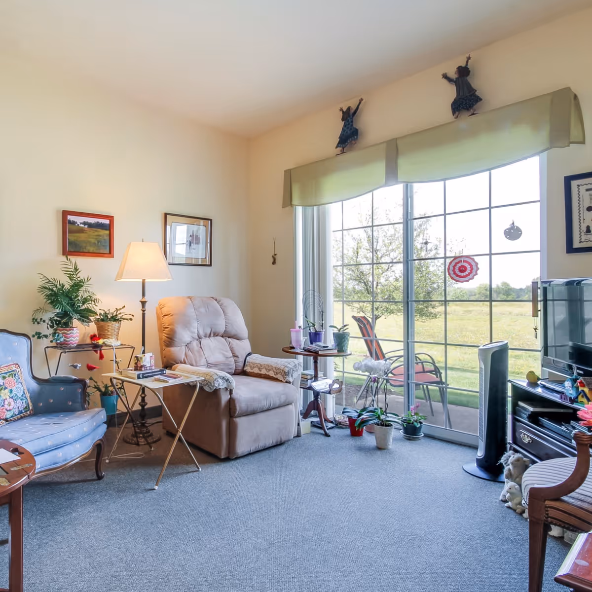 A cozy living room with a beige recliner chair, a blue upholstered armchair with a floral pillow, a floor lamp, and several potted plants. There is a large sliding glass door with a view of a grassy outdoor area and two patio chairs. The room has light-colored walls with framed pictures and decorative figurines above the window. A TV and fan are positioned on the right side of the room.