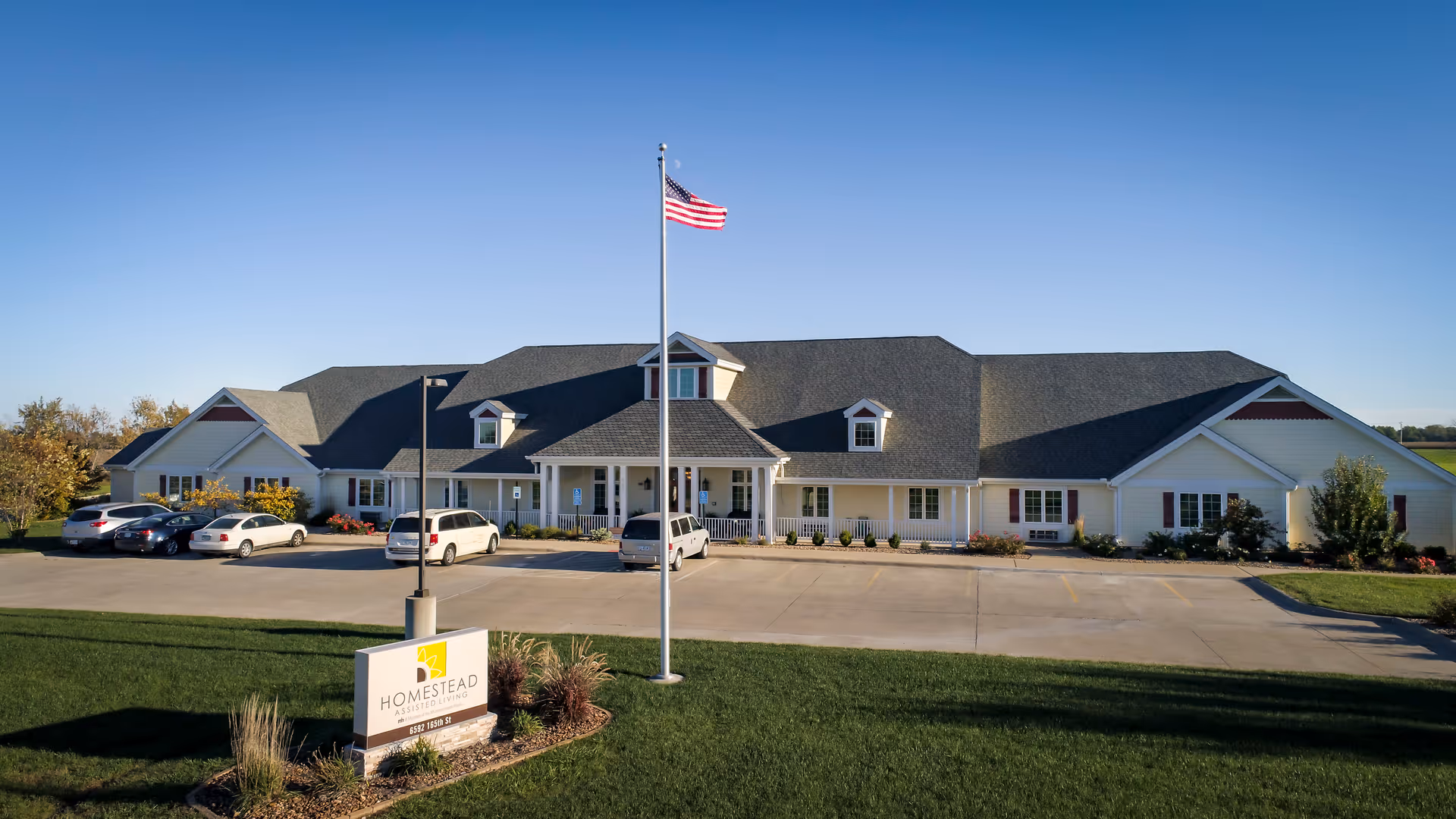 Exterior view of Homestead Assisted Living of Albia, a single-story building with a dark gray roof and white siding. There is a parking lot with several cars in front, a flagpole with an American flag, and a sign displaying the facility's name near the entrance. The sky is clear and blue.