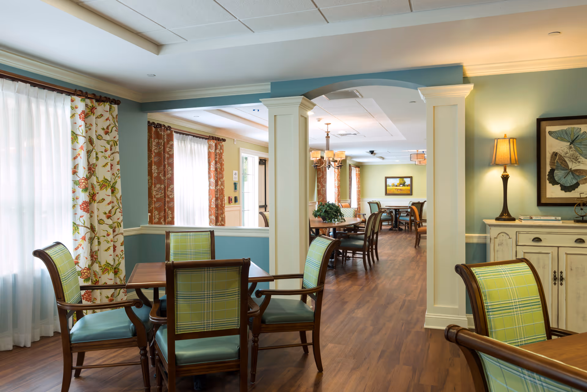 Communal dining room with wooden tables and green plaid upholstered chairs, warm lighting, and large windows.
