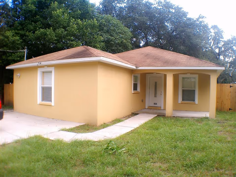 Single-story yellow building with a brown shingled roof, two windows with white frames, a white front door with the number 908, a concrete walkway leading to the entrance, and green grass in the front yard surrounded by trees.
