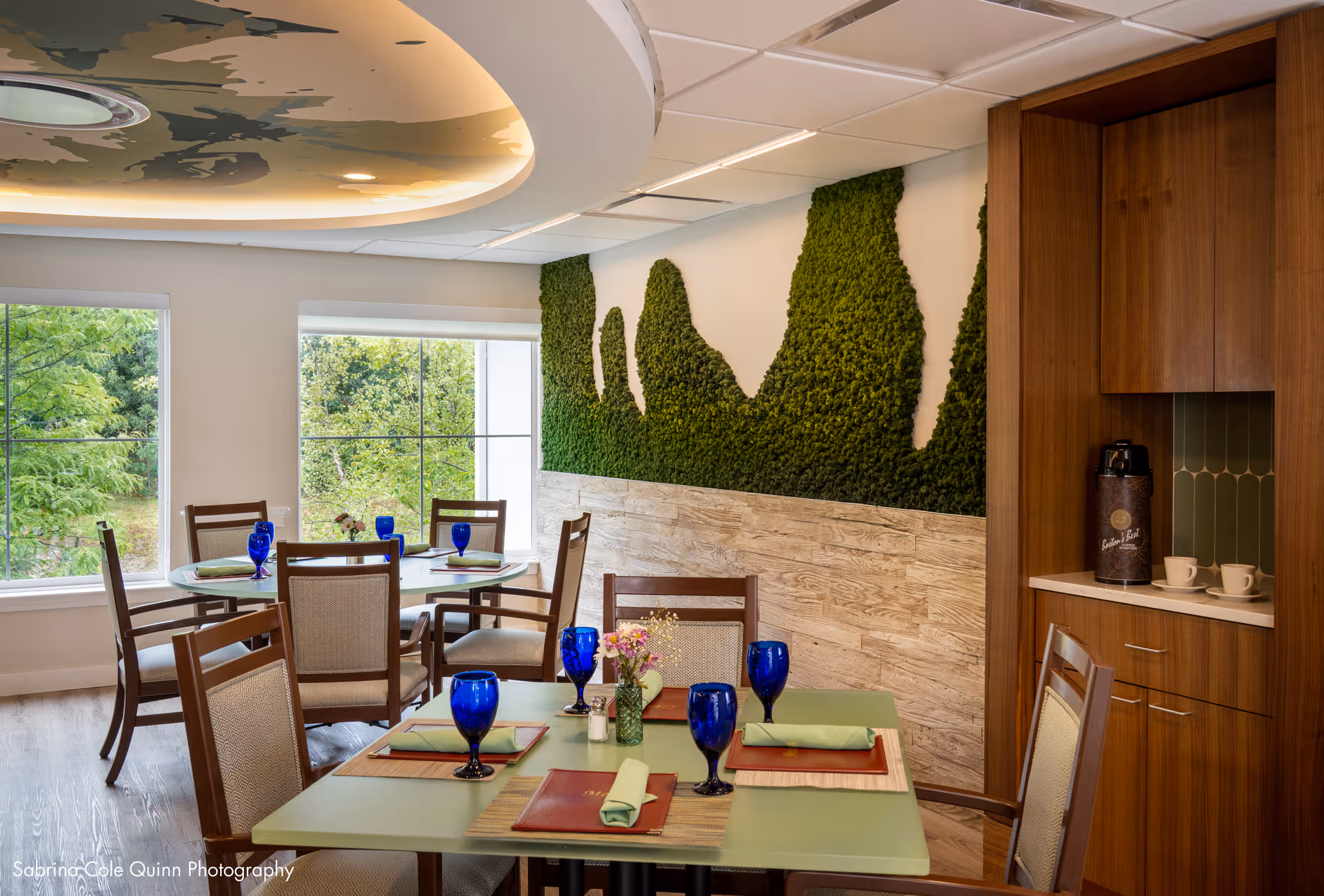 Bright dining room with moss wall art, round tables set with blue glasses and windows.