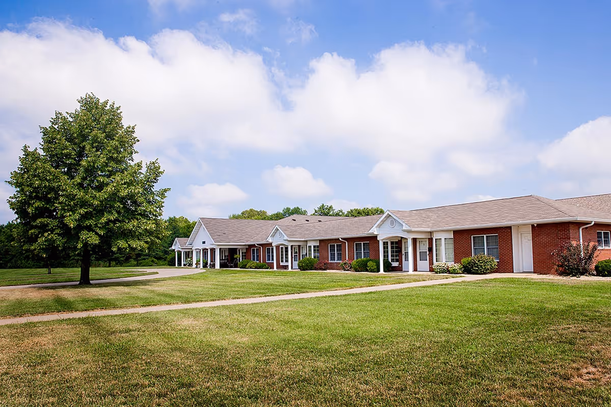 A single-story brick building with white trim and multiple windows, surrounded by a well-maintained green lawn and a large tree under a partly cloudy blue sky.