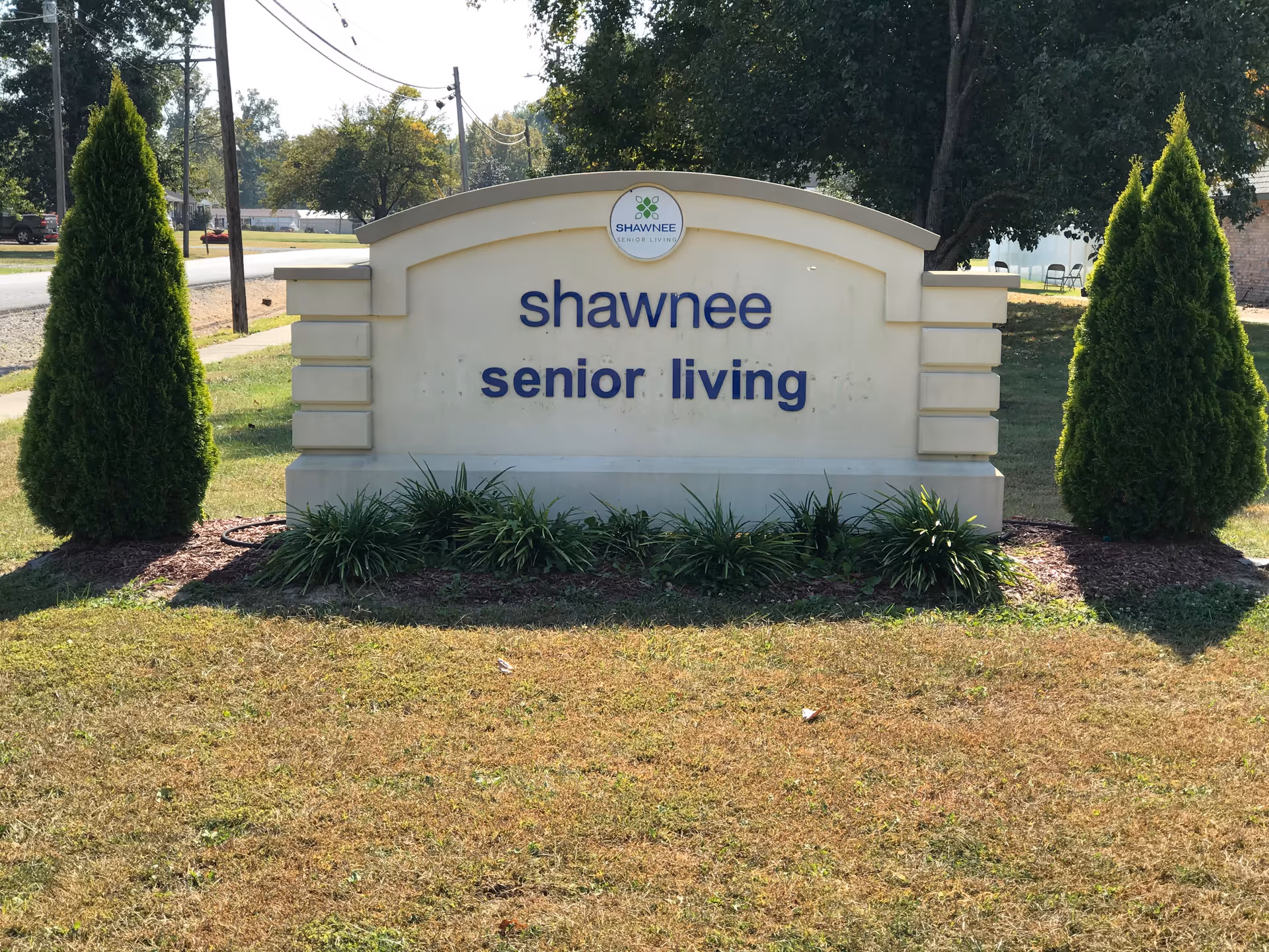 Outdoor sign for Shawnee Senior Living facility, surrounded by grass and two tall evergreen shrubs on either side, with trees and a road in the background.