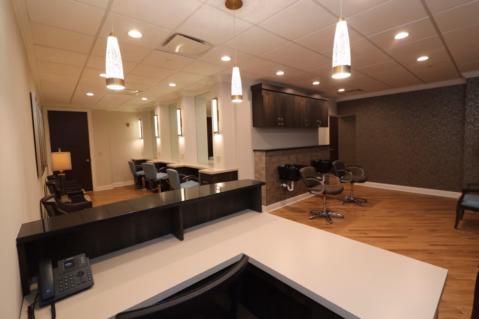Interior view of a salon area in a senior living facility with a reception desk in the foreground, salon chairs and hair washing stations along the right wall, and styling stations with mirrors and chairs along the left wall. The room has wooden flooring, pendant lights, and a patterned accent wall.