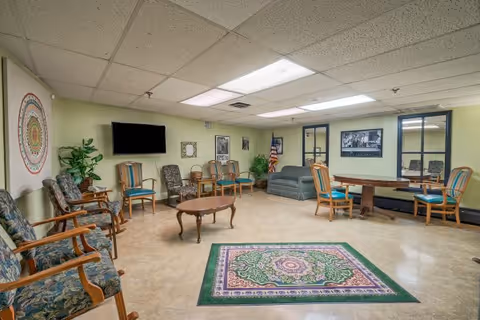 A spacious common room with multiple wooden chairs arranged along the walls and around a wooden coffee table. The room features a patterned area rug in the center, a small sofa against the far wall, an American flag, and framed pictures on the walls. There is a flat-screen TV mounted on one wall and fluorescent ceiling lights illuminating the room.