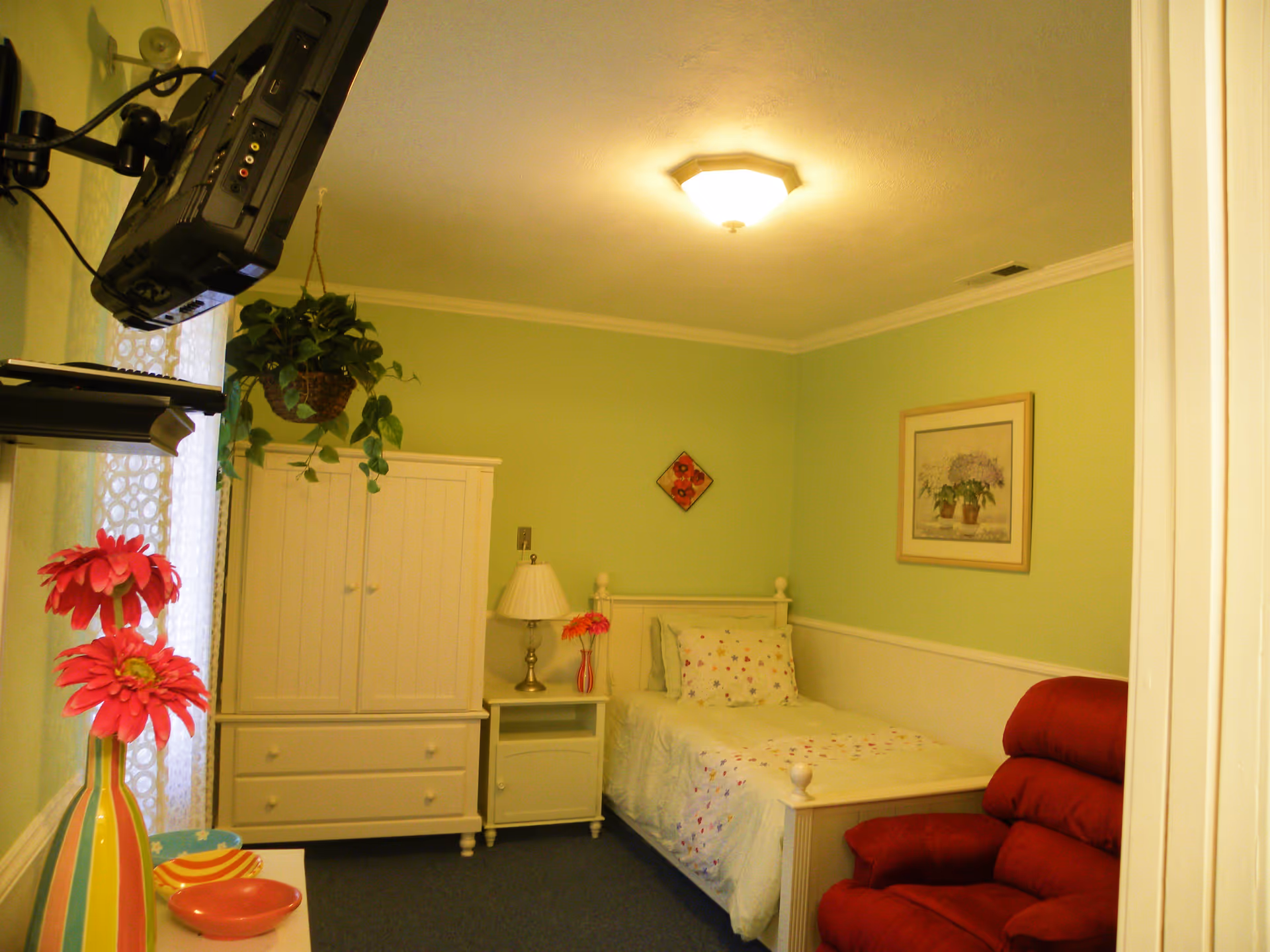 Small single bedroom with light green walls, a white bed and wardrobe, a red recliner, and a wall-mounted TV.