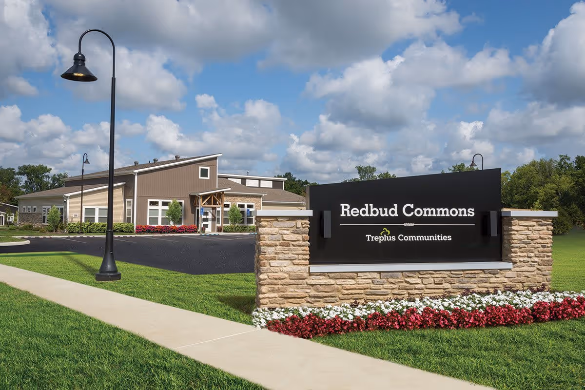 Entrance sign reading 'Redbud Commons' in front of a low-slung community building with a manicured lawn, sidewalk, and lampposts under a partly cloudy sky.