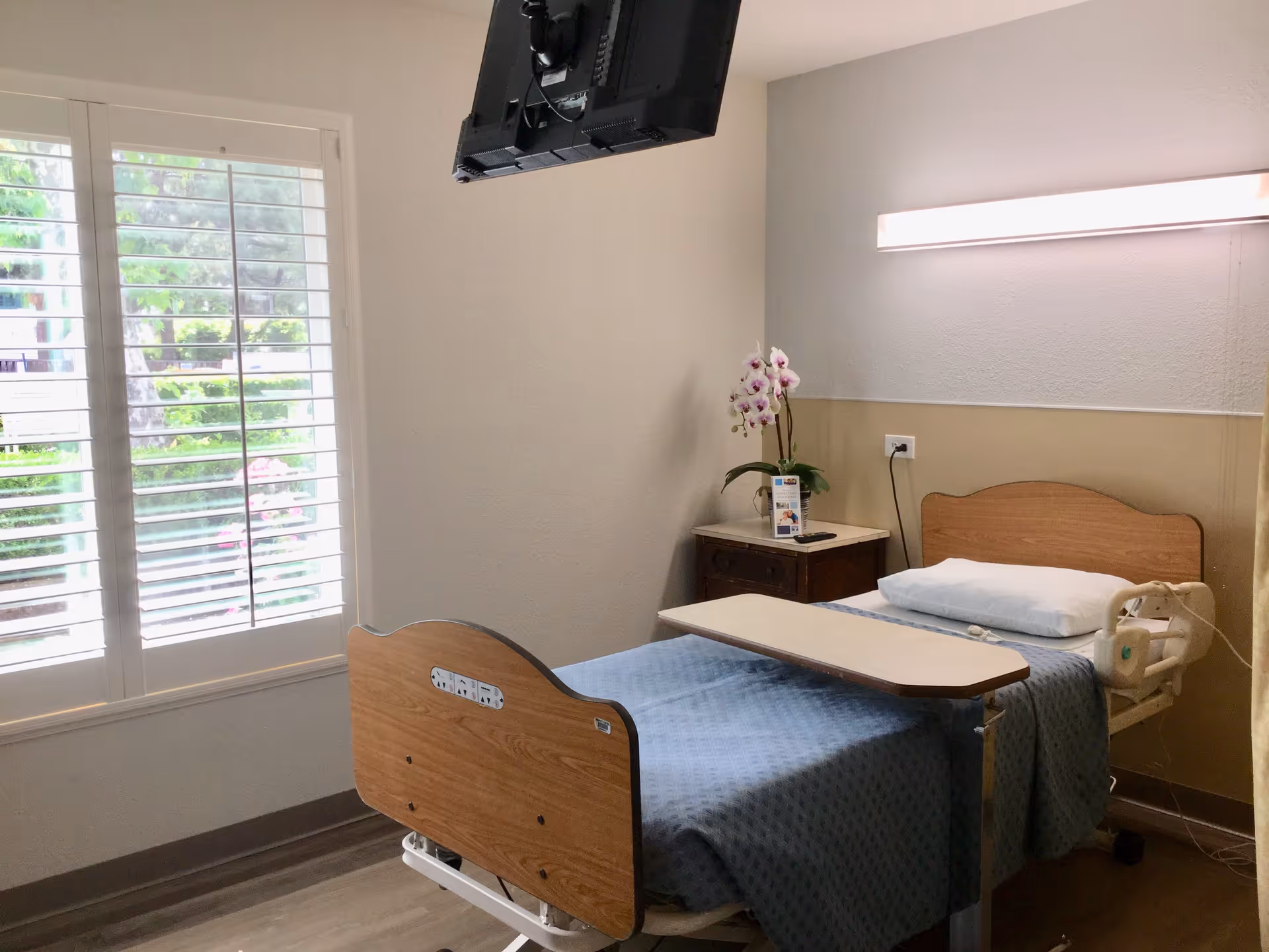 A clean and simple patient room in a skilled nursing facility featuring a single hospital bed with a blue blanket and white pillow. The bed has an overbed table positioned on top. Next to the bed is a small wooden nightstand with a potted orchid and informational pamphlets. A flat-screen TV is mounted on the wall above the bed. The room has a large window with white shutters letting in natural light and a beige wall with a horizontal light fixture above the bed.