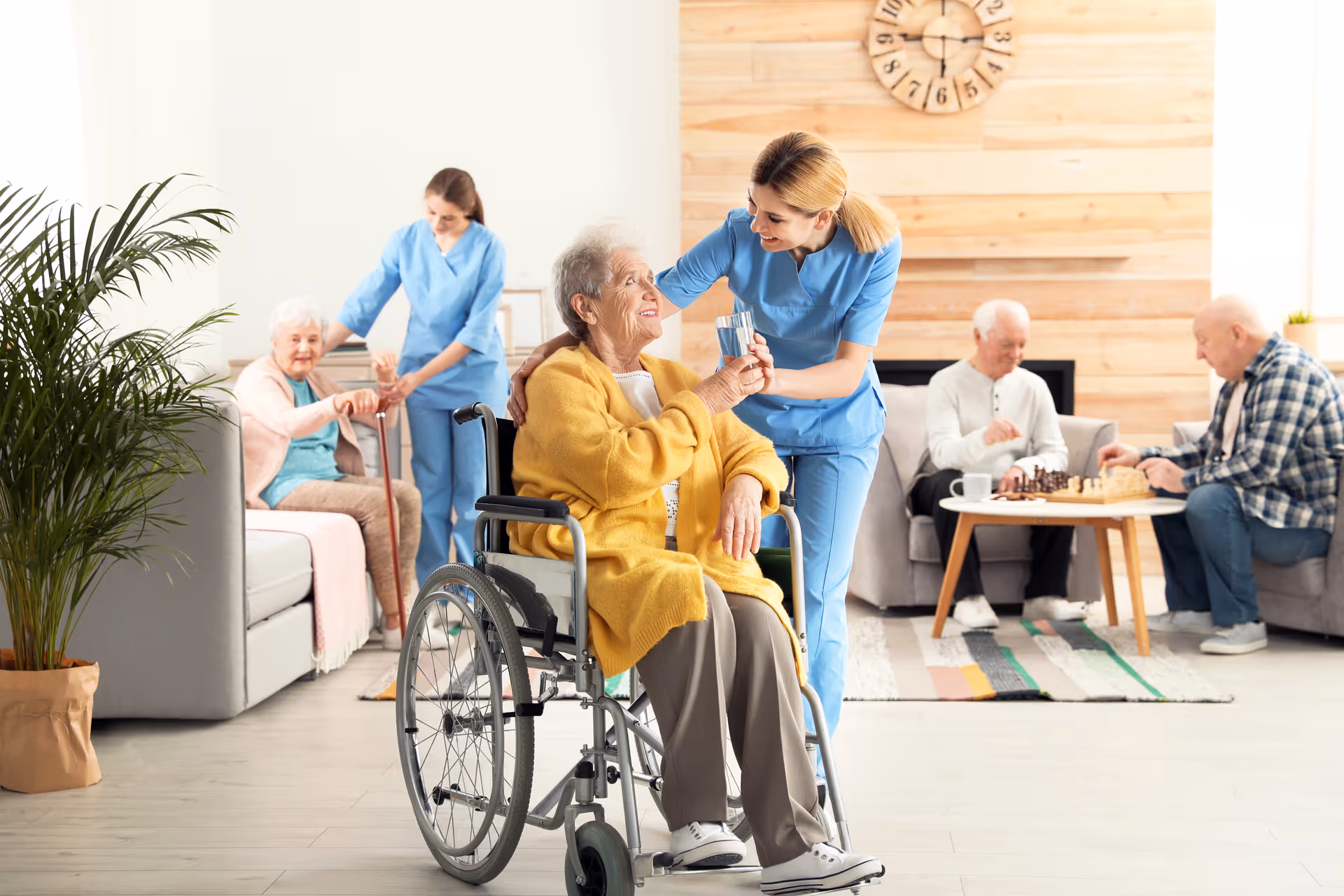 A senior woman in a yellow cardigan sitting in a wheelchair is being handed a glass of water by a smiling caregiver in blue scrubs in a bright, cozy living room. In the background, another caregiver assists an elderly woman sitting on a couch, while two elderly men play chess at a small table. The room has light-colored walls, a large clock on a wooden accent wall, and a potted plant.
